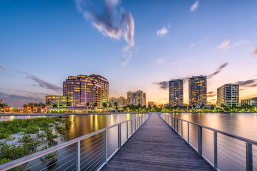 Sunset view of a modern waterfront skyline in West Palm Beach, Florida, with a wooden pier leading toward high-rise buildings, symbolizing urban growth and real estate investment potential in South Florida.